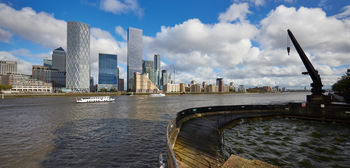 Canary Wharf Dock Crane 1 This urban landscape photograph, taken in the afternoon during early autumn, features Canary Wharf Dock Crane 1 prominently situated along the River Thames in London, United Kingdom. The image captures the historic dockside crane to the right, with the modern skyline of Canary Wharf rising across the water. Landmark skyscrapers, including the HSBC building and distinctive contemporary towers, are clearly visible in the background, illustrating the business district's architectural diversity. The river itself is central to the composition, with boats moving across its surface under a sky filled with clouds, encapsulating the dynamic urban environment of this area.